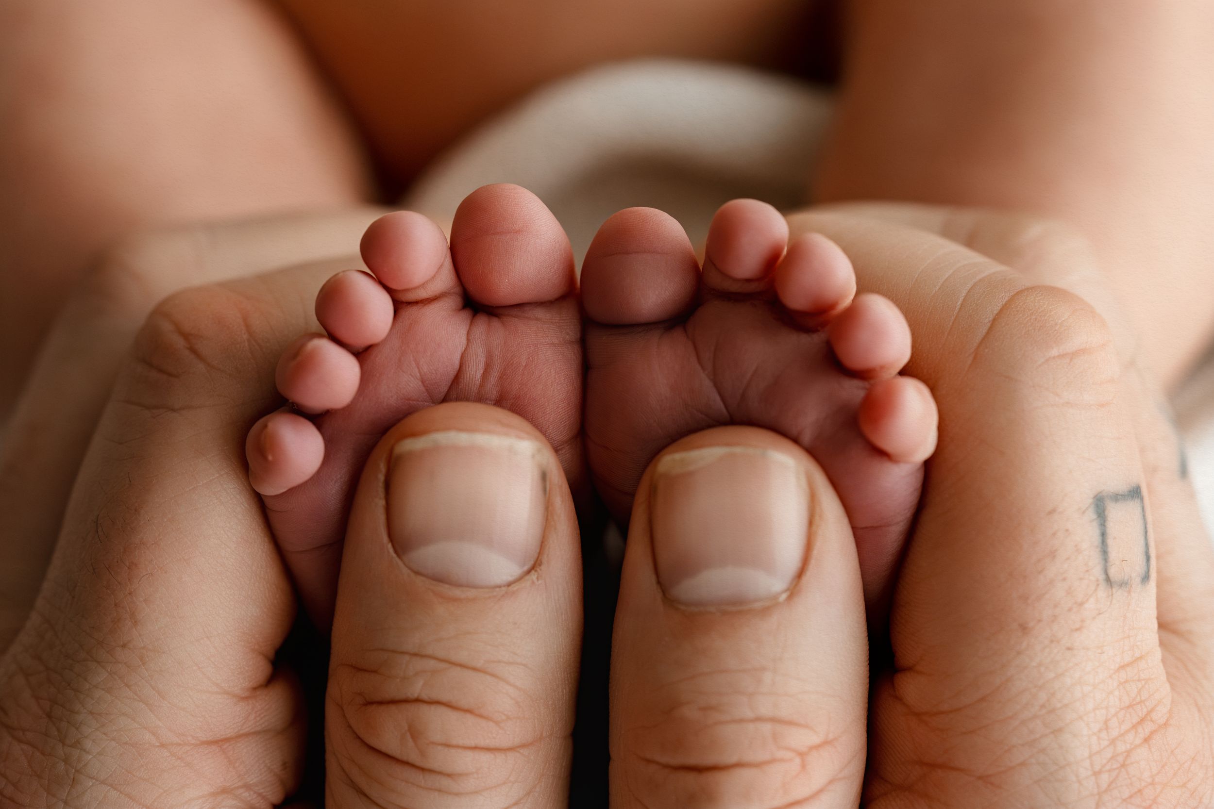 Close-up of tiny newborn baby feet cradled gently in adult hands with a small tattoo visible.