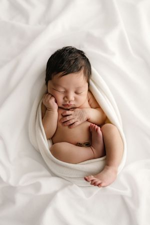 Peaceful newborn baby sleeping wrapped in white cloth on soft white blanket with dark hair and tiny hands tucked under cheek.