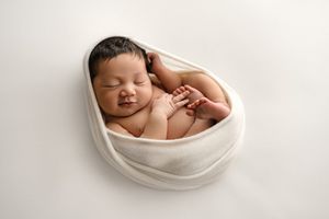Peaceful newborn baby wrapped in white cloth, sleeping curled up in a womb-like pose on a white background.