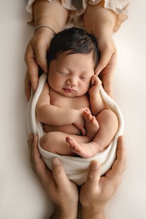 Newborn baby sleeping peacefully wrapped in white cloth, cradled gently in parents hands on soft background.