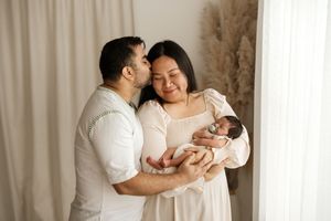 A couple in white outfits tenderly holds their newborn baby, with the father kissing the mother's cheek.