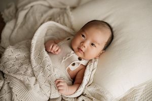 Adorable newborn baby wrapped in a cozy knit blanket, lying on a white bed, gazing up with bright eyes.