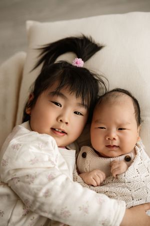 Young Asian girl with pink flower hair clip gently holds newborn baby sibling, both resting on cream cushion.