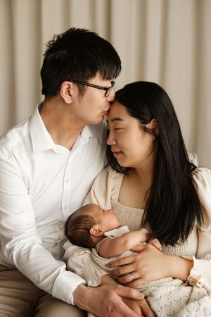 A couple tenderly holds their newborn baby, with the man kissing the woman's forehead in a warm family portrait.
