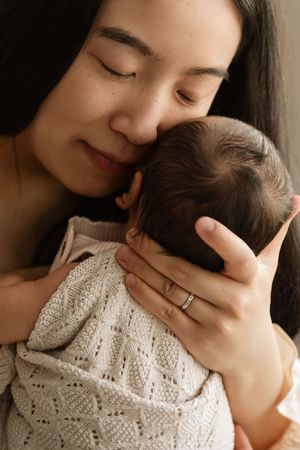 A tender moment as a young Asian mother gently nuzzles her sleeping newborn baby wrapped in a cream knit blanket.