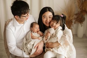 Happy Asian family portrait with parents holding newborn baby and toddler daughter in white outfits.