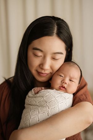 A young Asian mother tenderly holds her sleeping newborn baby wrapped in a white knit swaddle blanket.