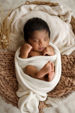 Peaceful newborn baby swaddled in white wrap sleeping in woven basket with cream knit blanket.