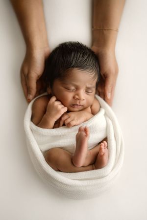 Newborn baby swaddled in white wrap sleeping peacefully while adult hands gently cradle its head from above.