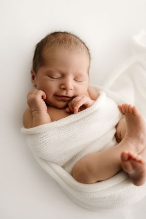 Sleeping newborn baby swaddled in white knit wrap with tiny hands near face on white background.