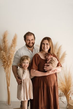 Family of four posing for newborn portrait with pampas grass decor, parents smiling with toddler daughter and newborn baby.