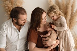Family portrait of parents and toddler girl lovingly gazing at newborn baby, with pampas grass decor backdrop.