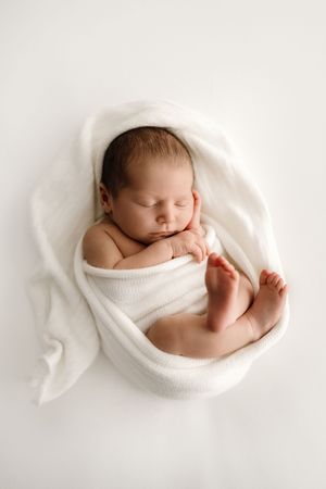 Peaceful newborn baby swaddled in white wrap sleeping curled up on white background.