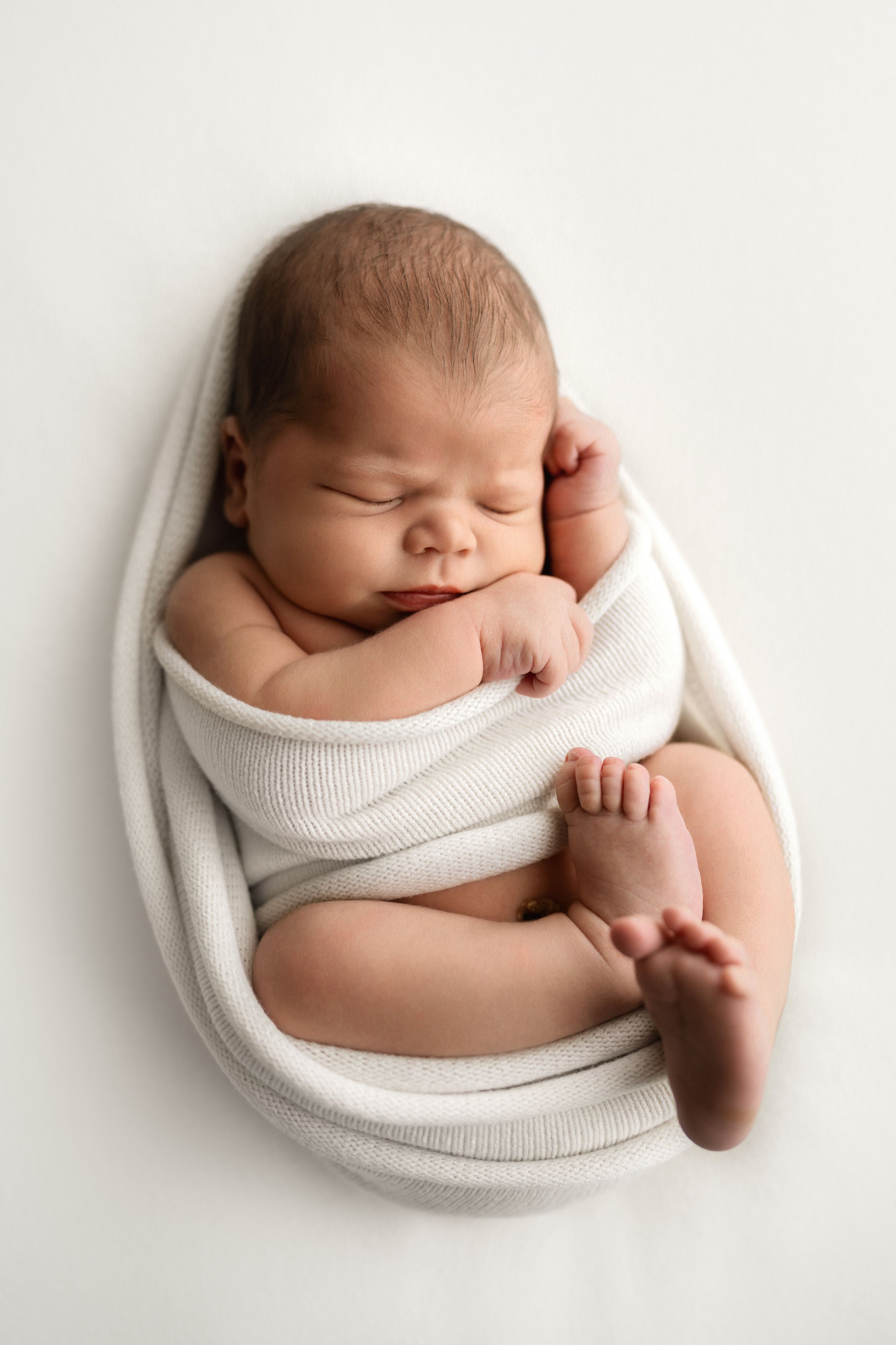 Peaceful newborn baby sleeping wrapped in a white knit blanket, curled in a natural fetal position on white background.