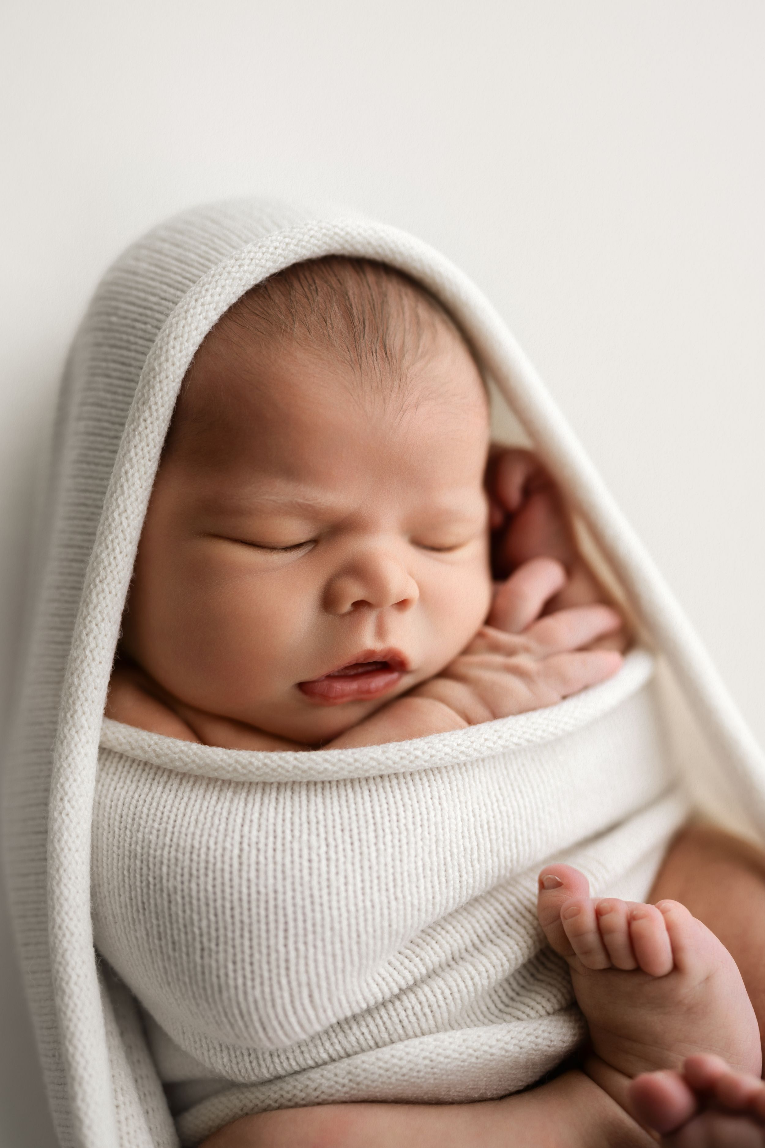Peaceful newborn baby sleeping wrapped in a soft white knit blanket with hood, eyes closed, tiny fingers visible.