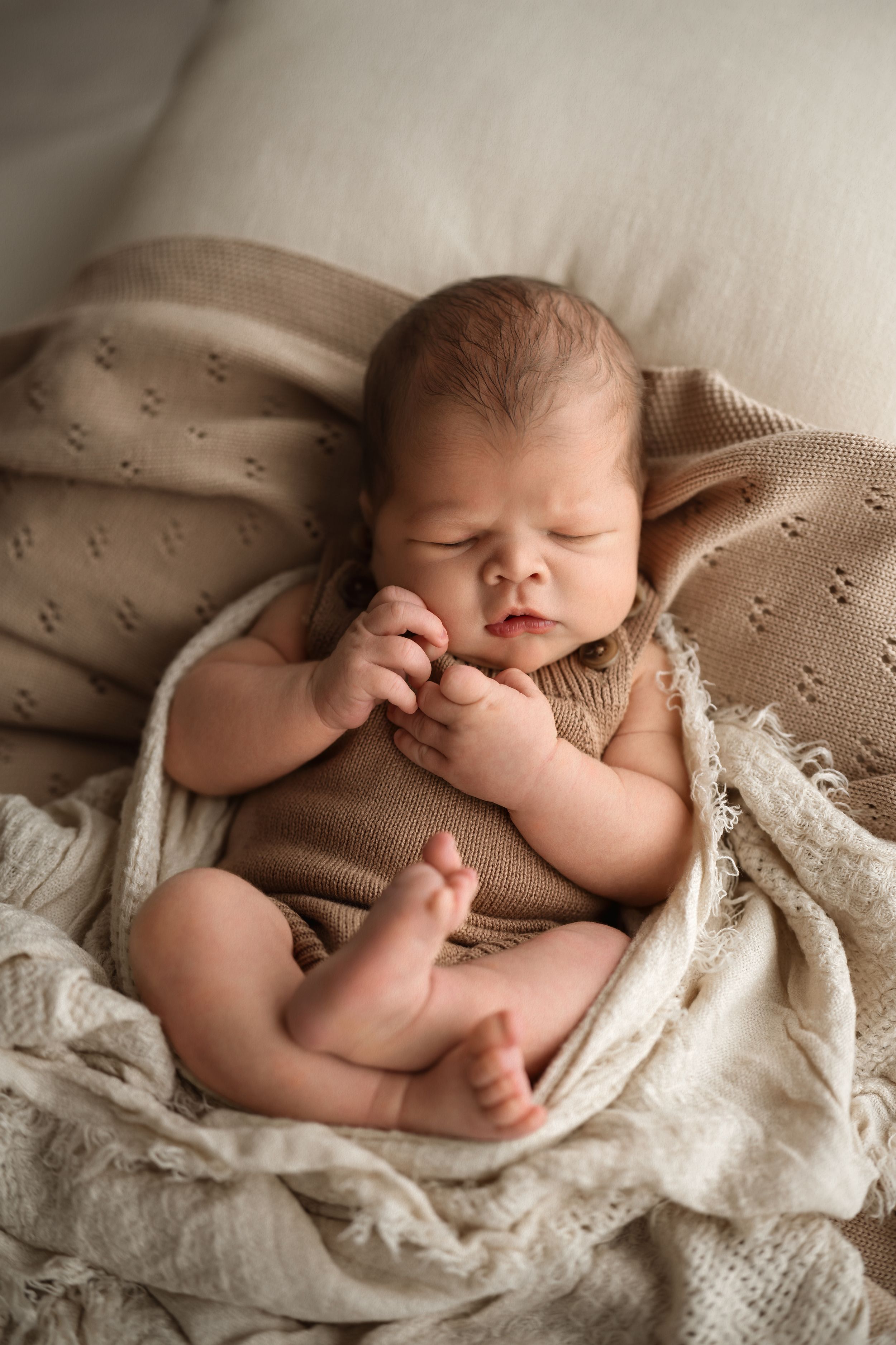 Peaceful newborn baby sleeping in beige knit romper, nestled in cream textured blankets, hands clasped together.