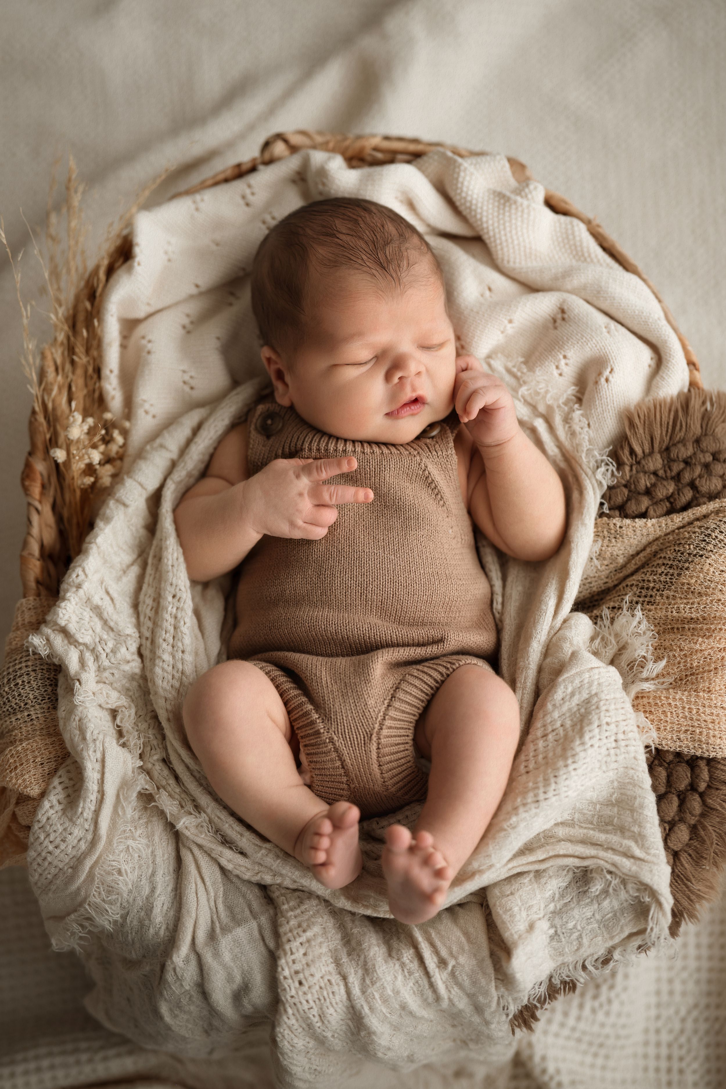 Sleeping newborn baby in knit brown romper nestled in wicker basket with cream blanket and dried wheat decor.