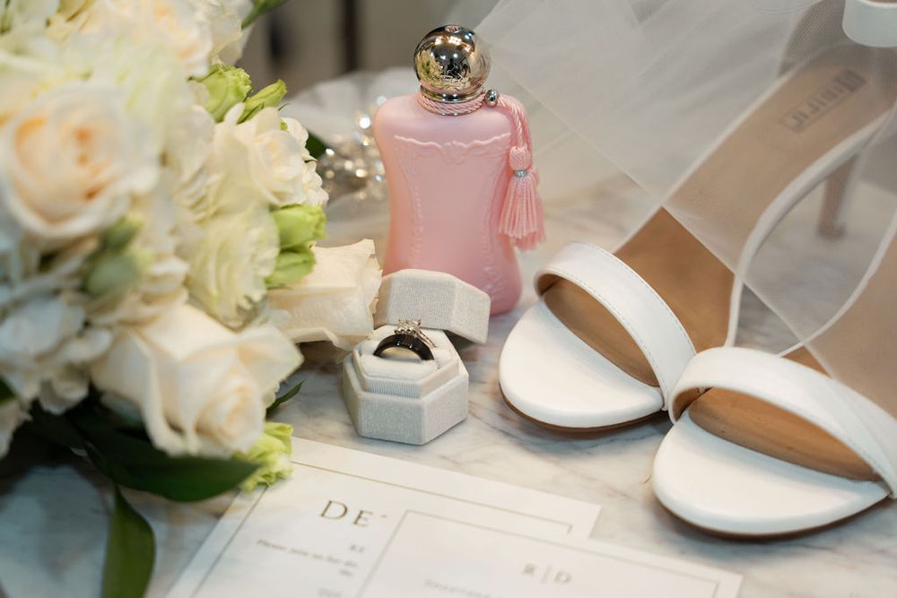 Wedding flat lay with white roses, diamond ring, pink perfume bottle, white heels, and monogrammed stationery.