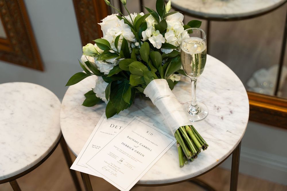 Elegant wedding bouquet of white roses and greenery beside champagne glass and invitation cards on marble table.