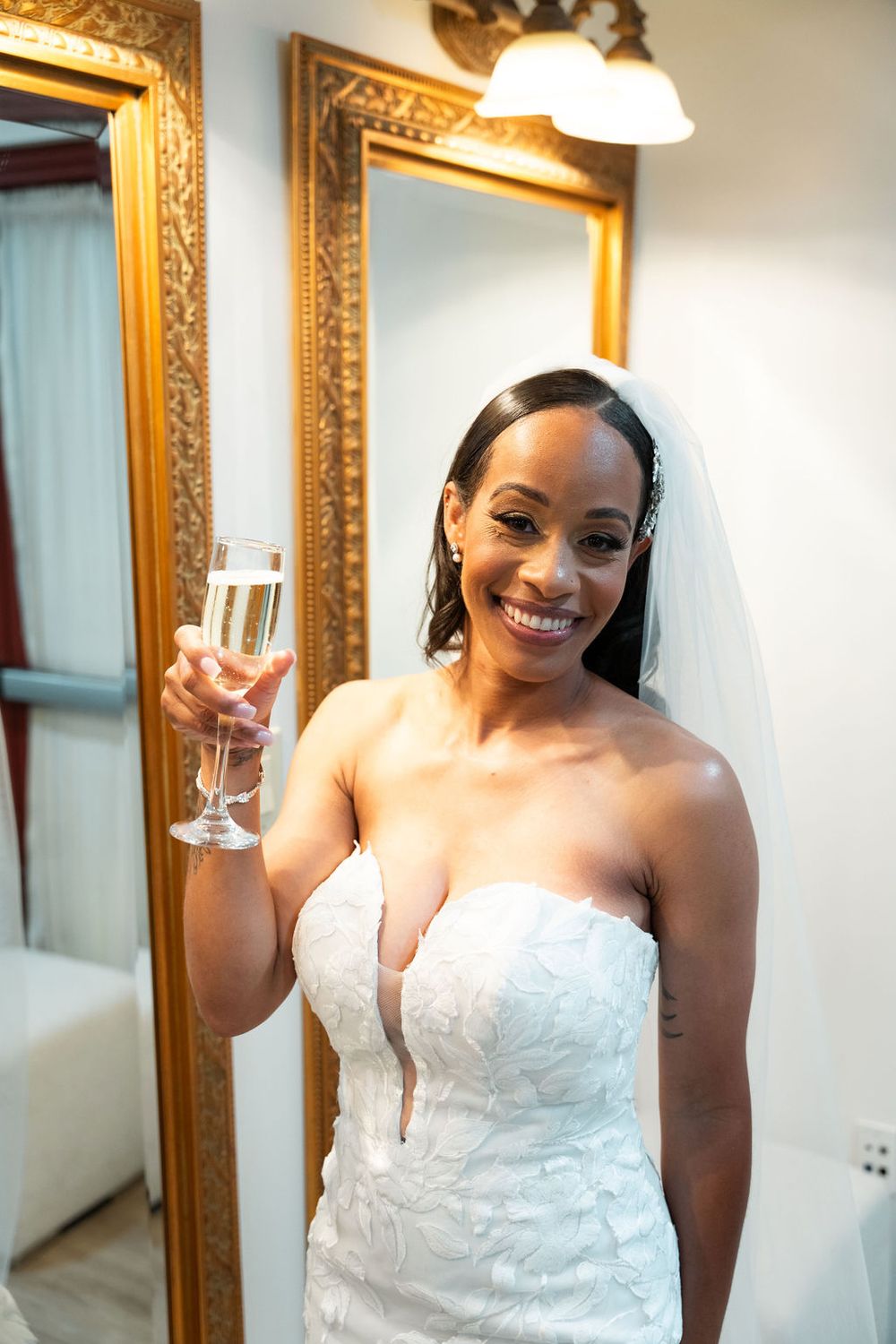 Smiling bride in white strapless gown and veil raises champagne glass in front of ornate gold mirror.