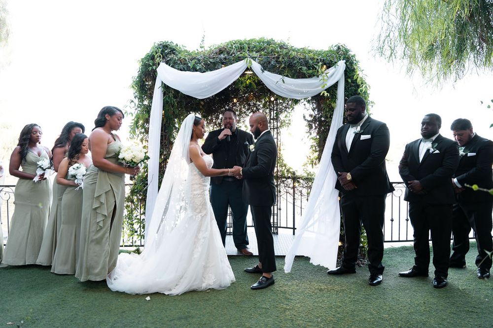 A bride and groom exchange vows under a floral arch, surrounded by bridesmaids in silver gowns and groomsmen in tuxedos.