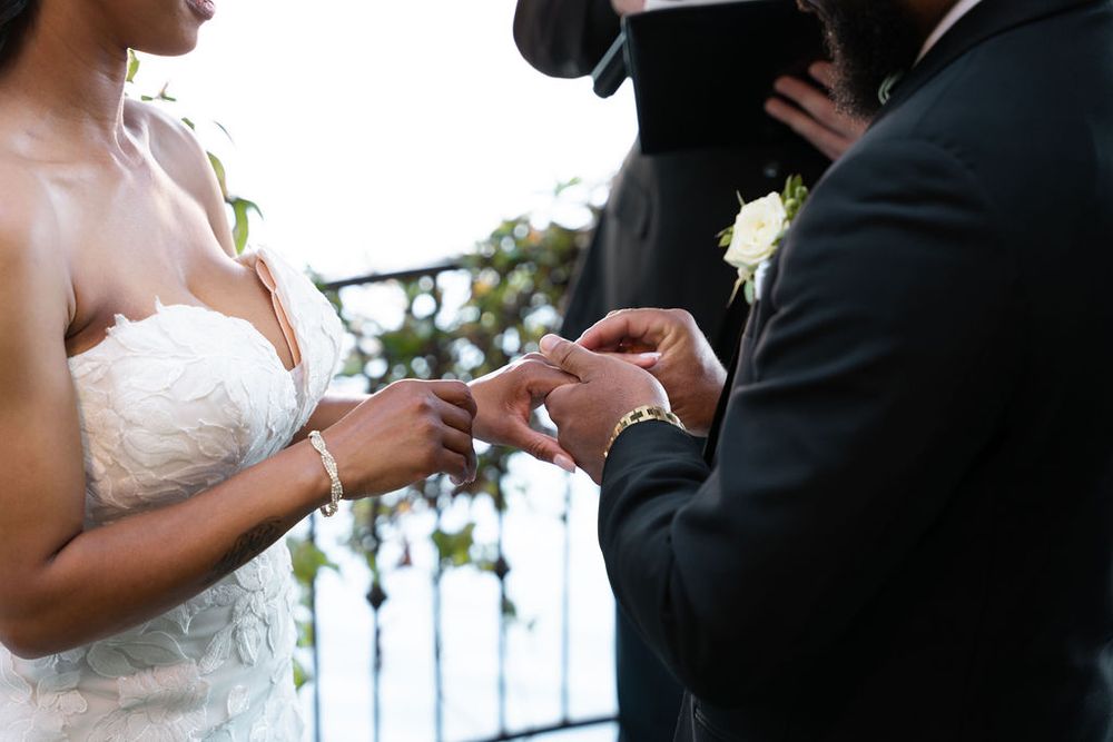 Bride and groom exchanging rings during an outdoor wedding ceremony, officiant wearing mask in background.