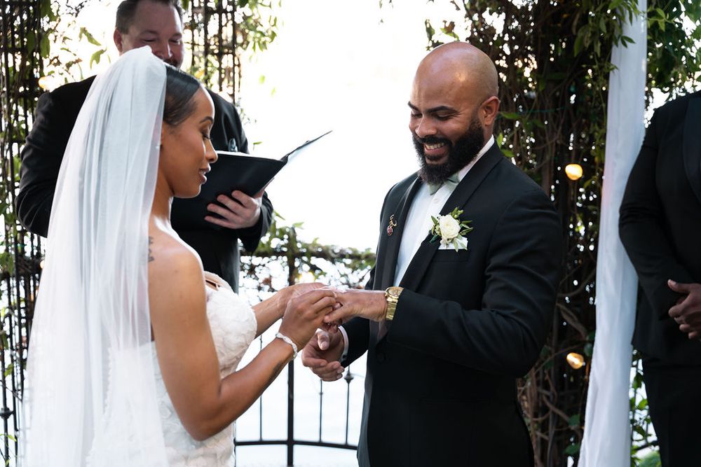 Groom places wedding ring on bride's finger during outdoor ceremony, both smiling joyfully.