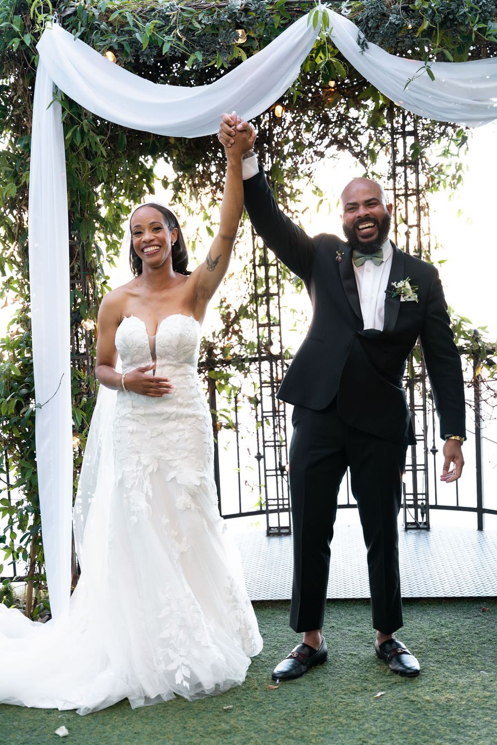 Joyful newlywed couple raising hands at outdoor wedding ceremony under white draped arch with greenery.