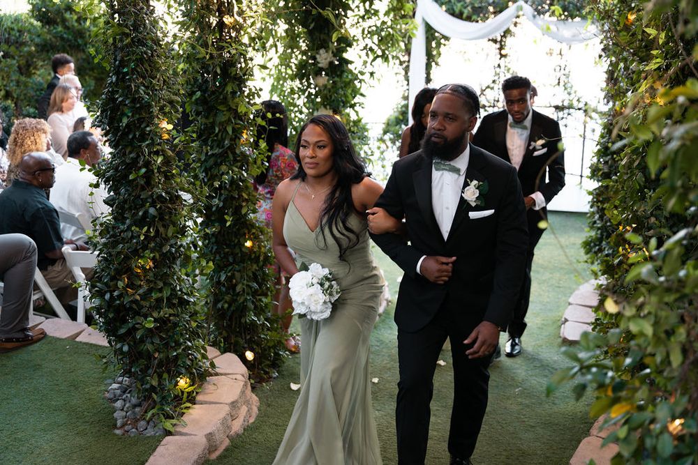 Bride in sage green gown walks with groom in black tuxedo down floral outdoor wedding aisle.