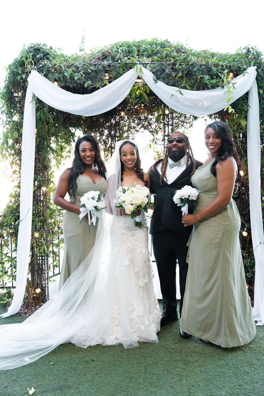 Bride in white gown with bridesmaids in sage green dresses and groom posing under floral arch at outdoor wedding.