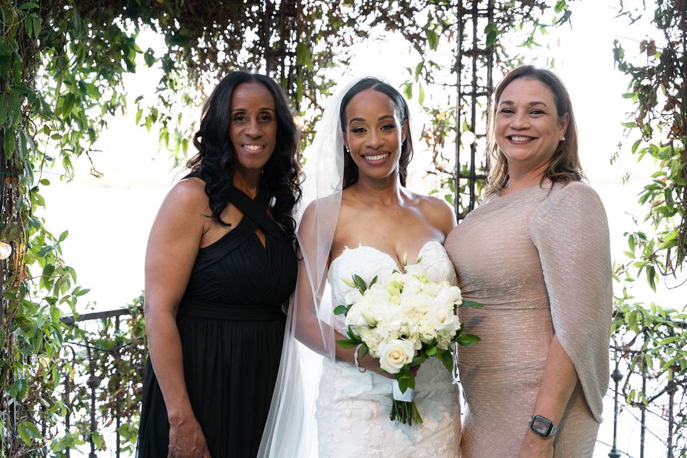 Bride in white lace gown and veil holds white bouquet, posing with two women at an outdoor wedding venue.
