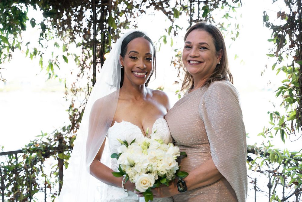 Bride in white gown holding bouquet poses with smiling woman in gold dress under floral archway.