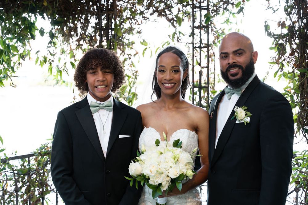 Bride in strapless gown holding white bouquet poses with two men in tuxedos at an outdoor wedding ceremony.