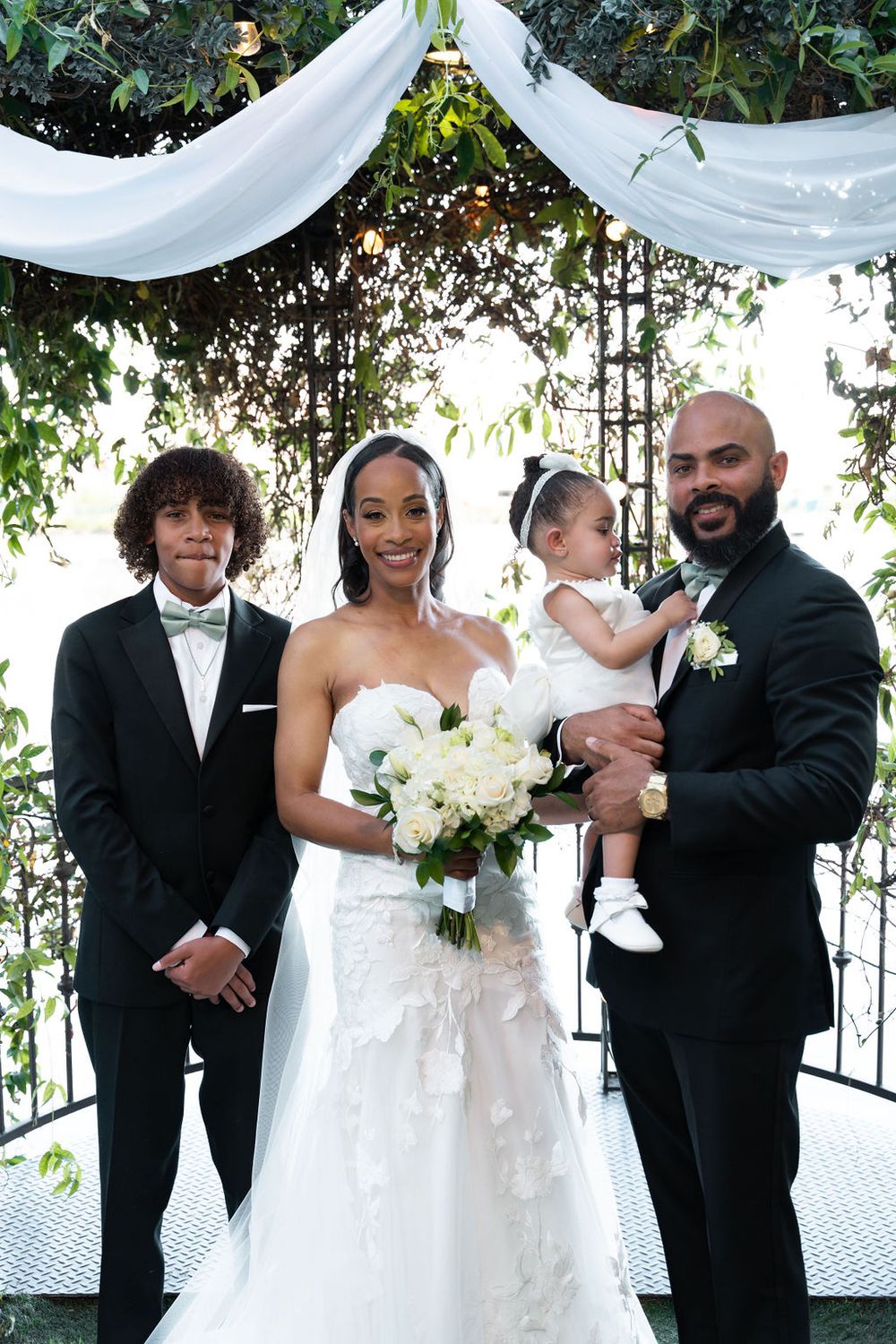 A bride and groom pose with their two children under a floral arch at an elegant outdoor wedding ceremony.