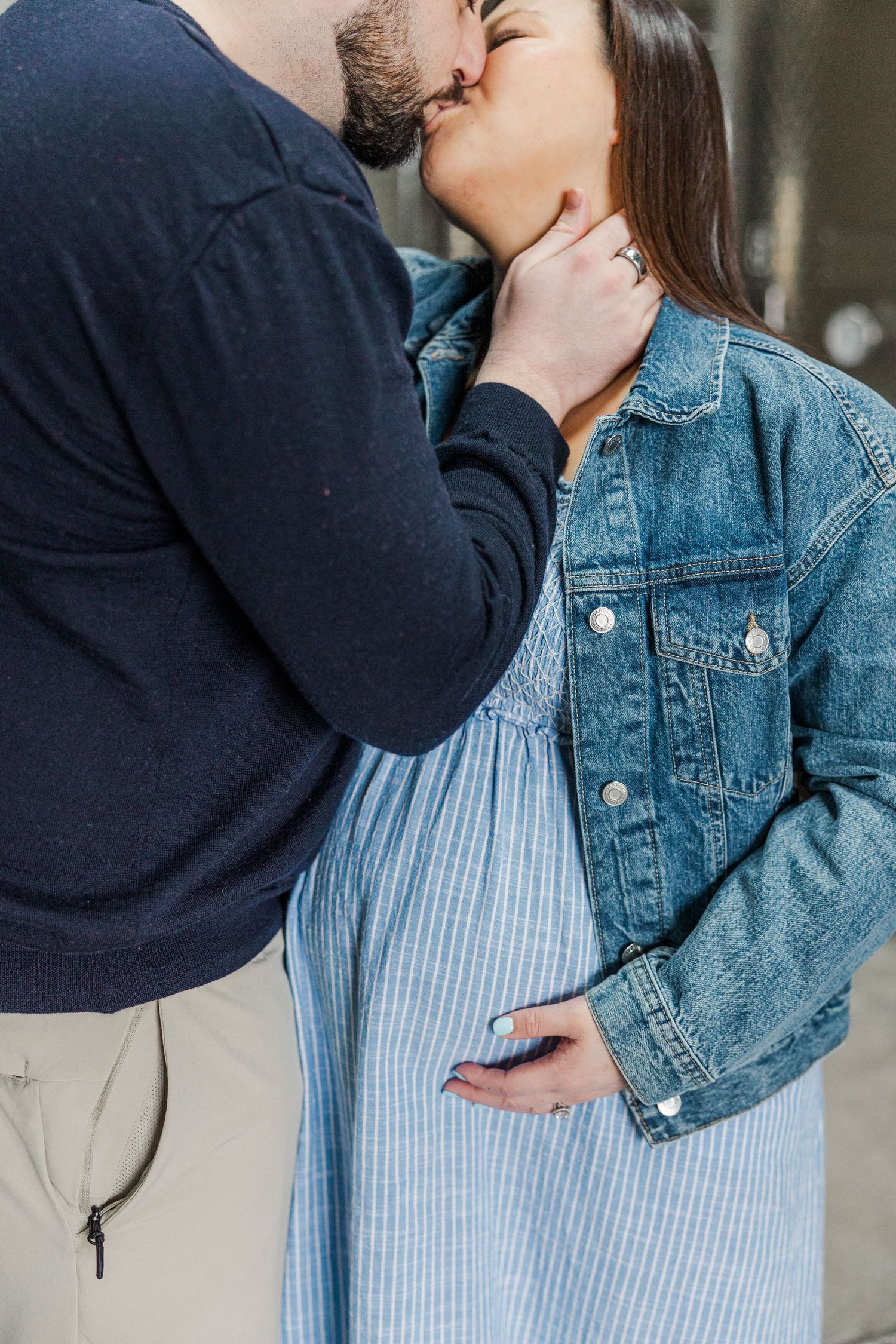 Pregnant couple sharing a tender kiss, woman in denim jacket and blue dress cradling her baby bump.