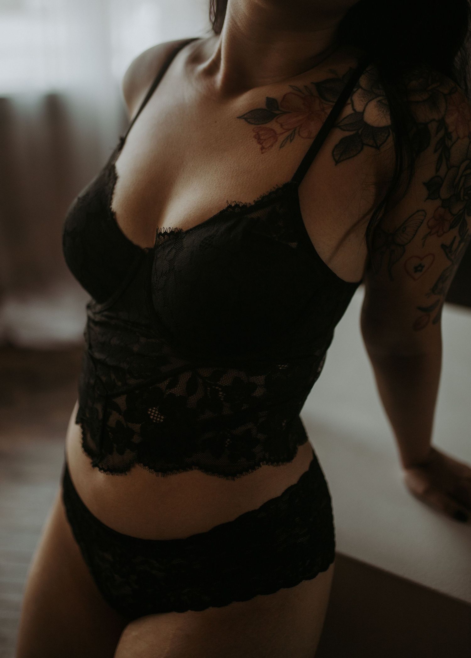 Woman in black lace lingerie set with floral tattoos on shoulder and chest posing in soft natural light.