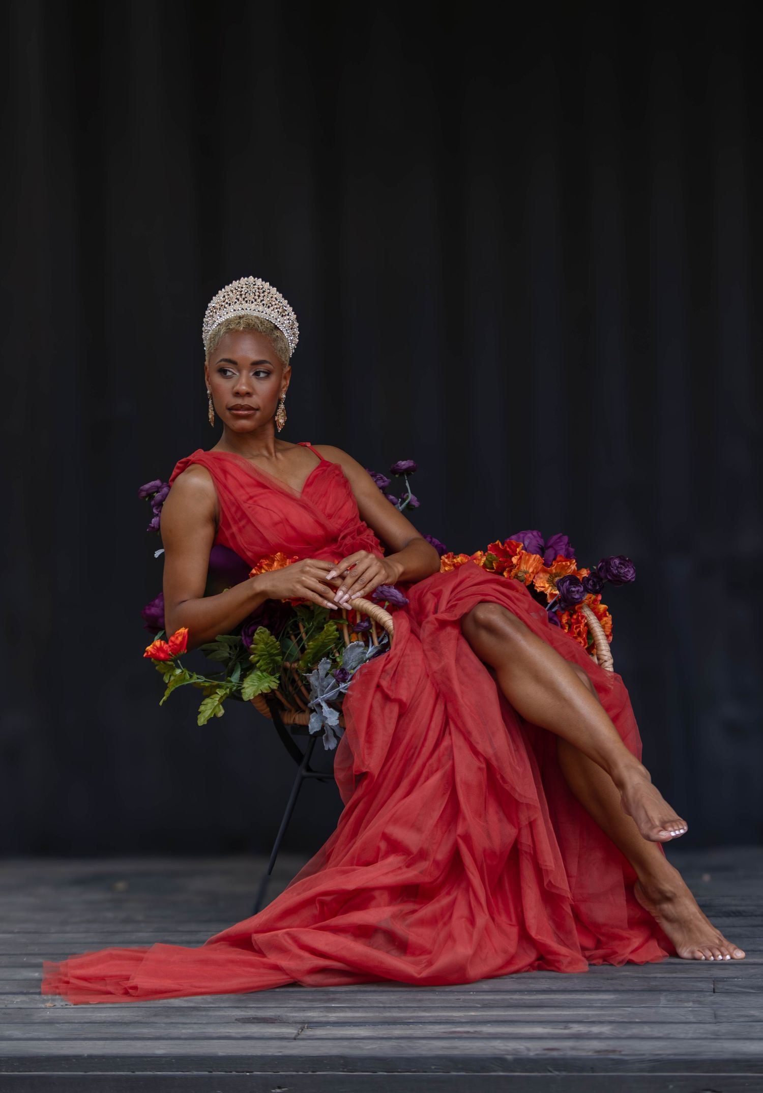 A Black woman in a red gown and pearl crown sits regally in a chair surrounded by colorful flowers.