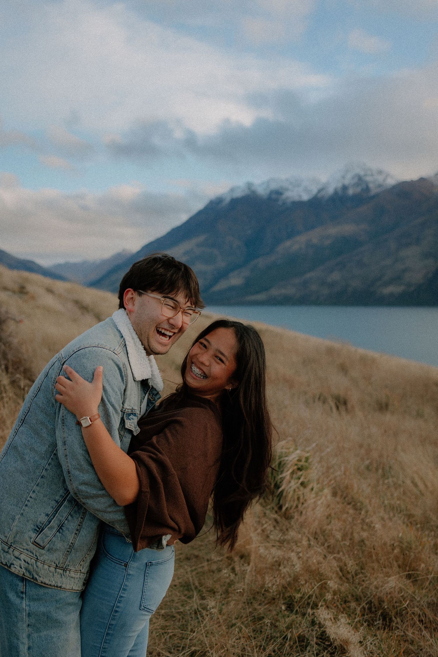 A joyful couple embraces and laughs together on a grassy hillside with a scenic lake and snow-capped mountains behind them.