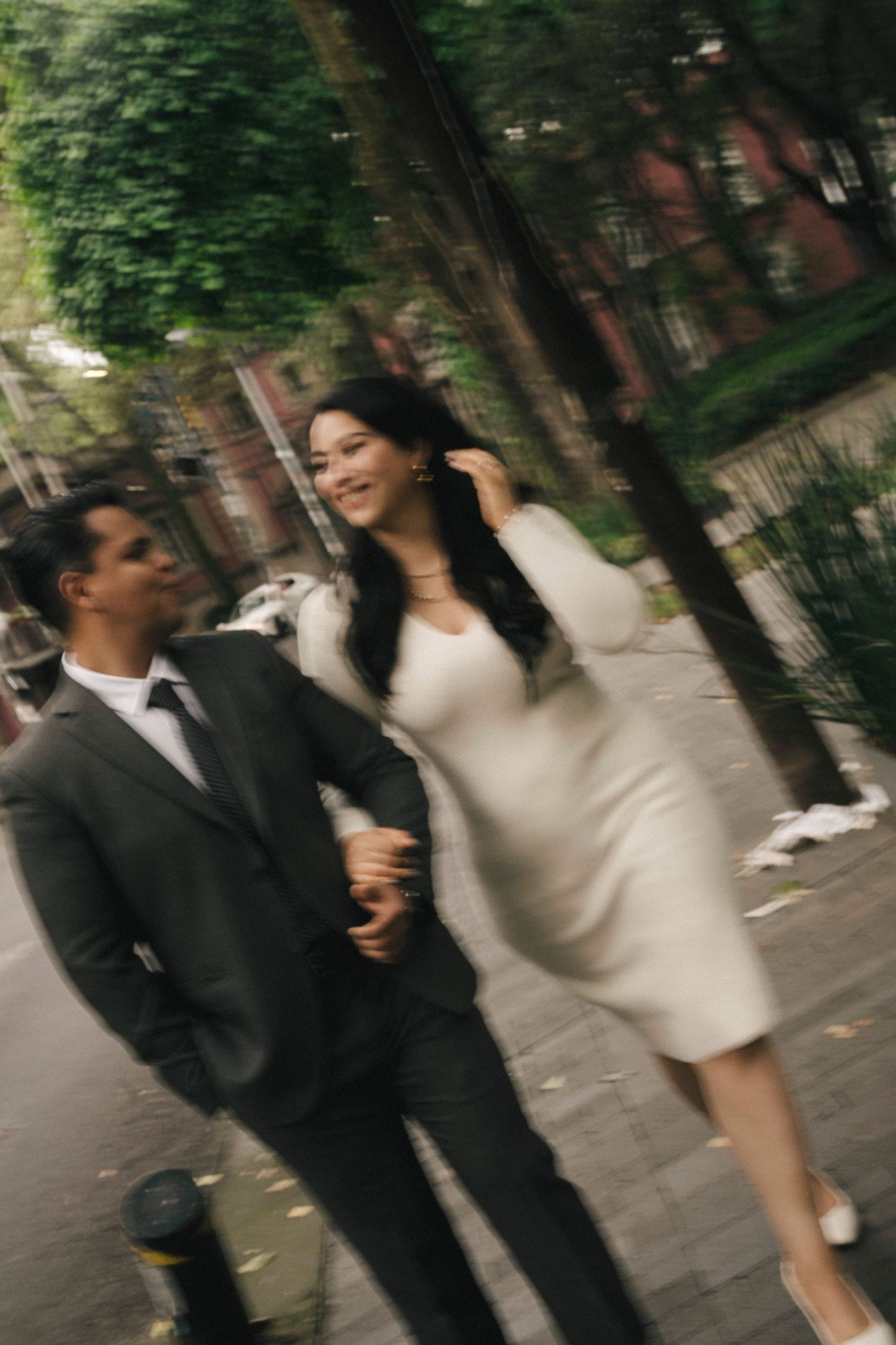 A couple in formal attire walks arm-in-arm down a tree-lined urban sidewalk, captured with motion blur.