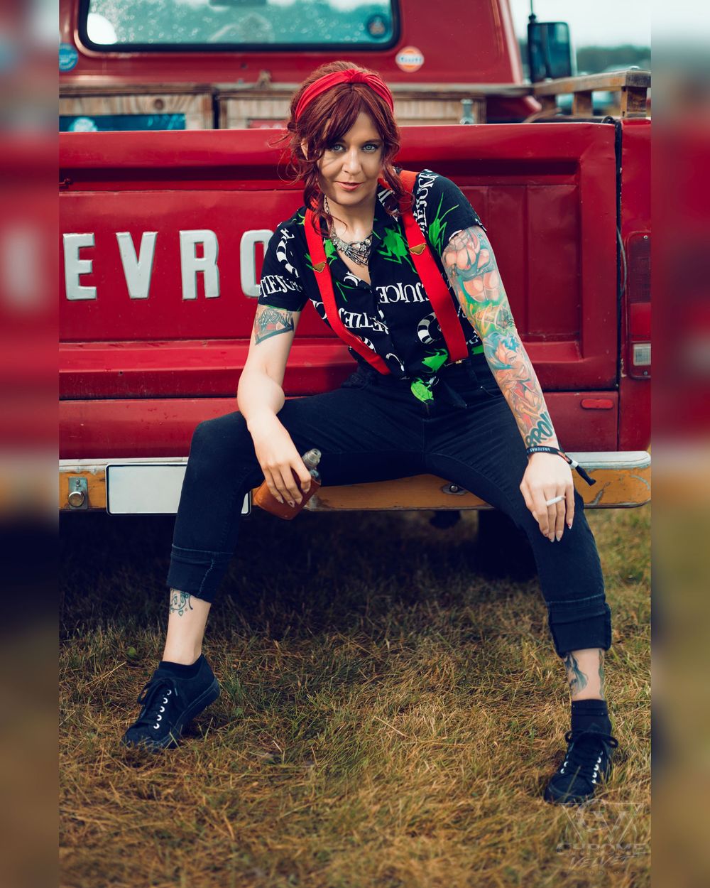 Tattooed woman with red hair in black outfit sitting on vintage red Chevrolet truck bumper outdoors.