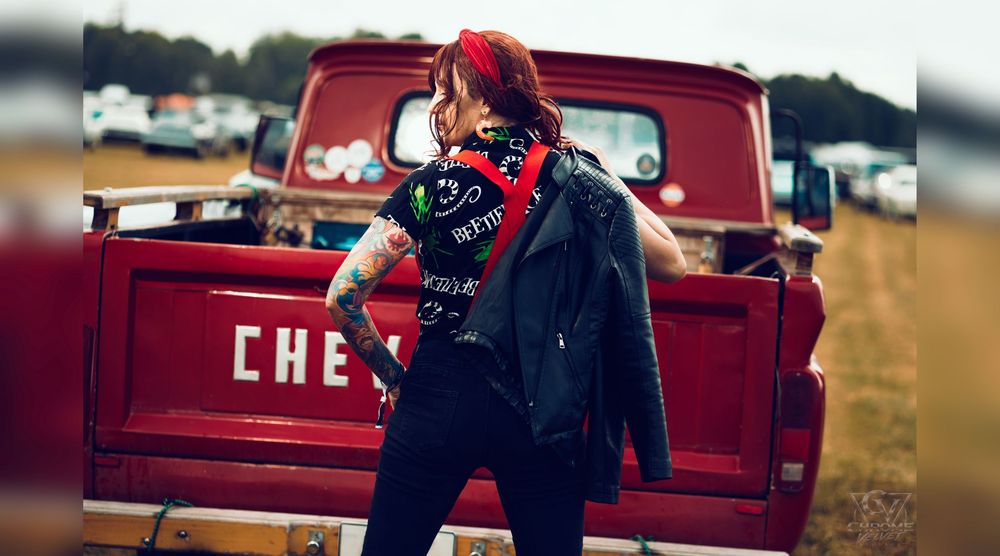 Tattooed woman in leather jacket poses near vintage red Chevy truck at outdoor festival.