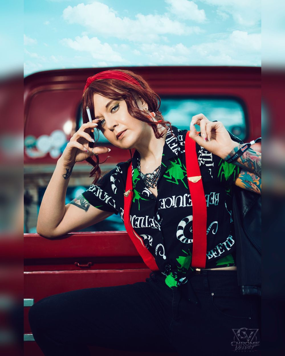 Young woman with red hair holding phone, wearing graphic tee with red suspenders, leaning against vintage red truck.