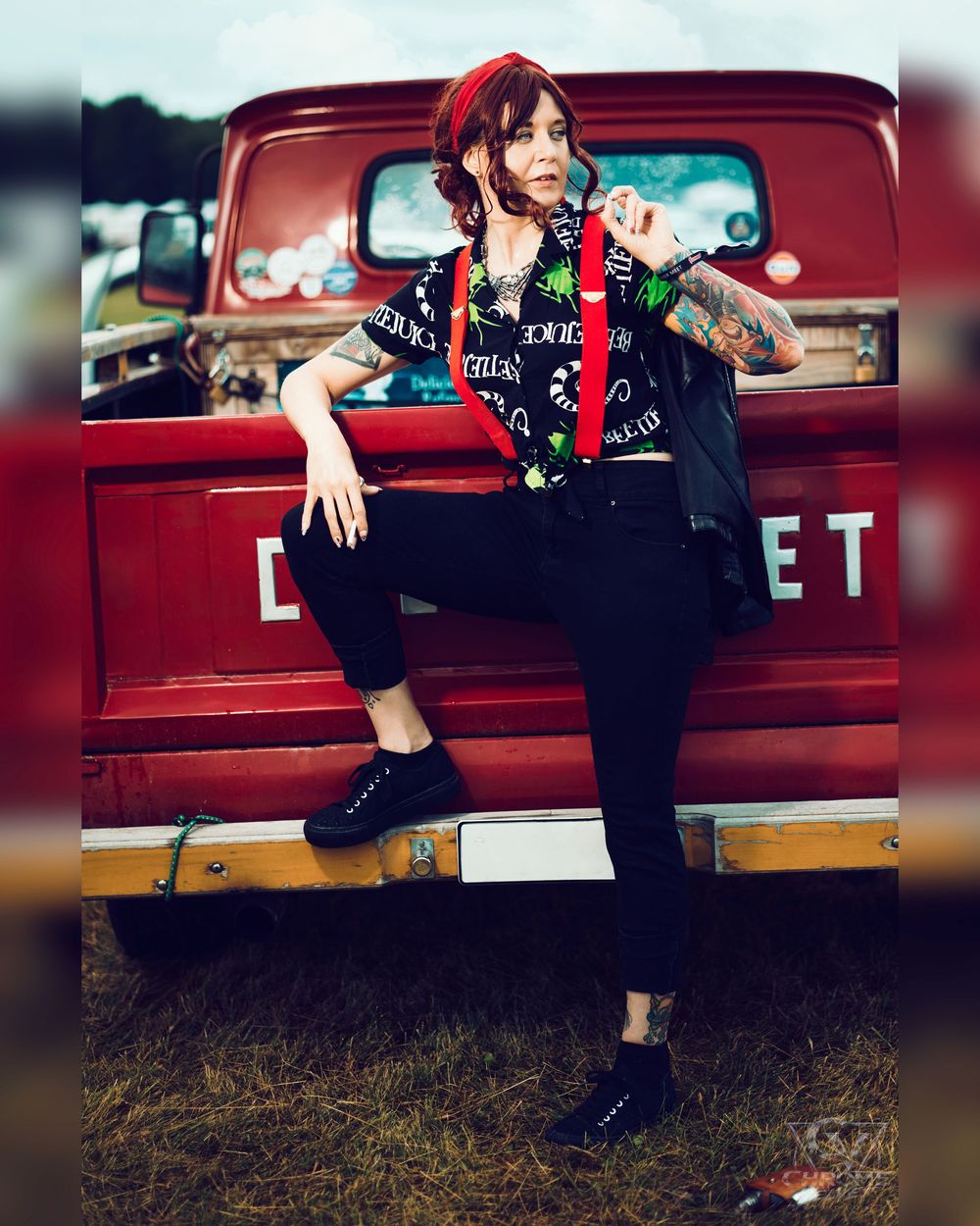 Tattooed woman in punk vest leans against vintage red Chevrolet truck outdoors.
