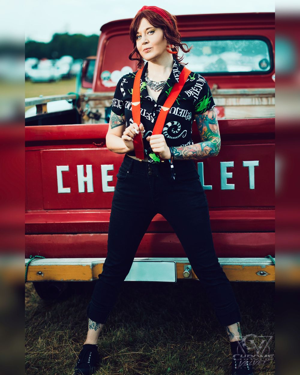 Tattooed woman in black outfit with red suspenders posing confidently in front of a vintage red Chevrolet truck.