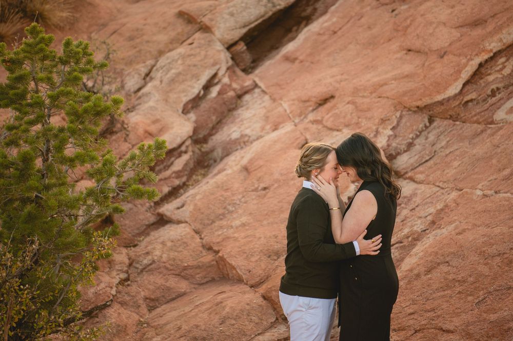 Two women share a romantic kiss in front of stunning red rock formations in an outdoor engagement photo.