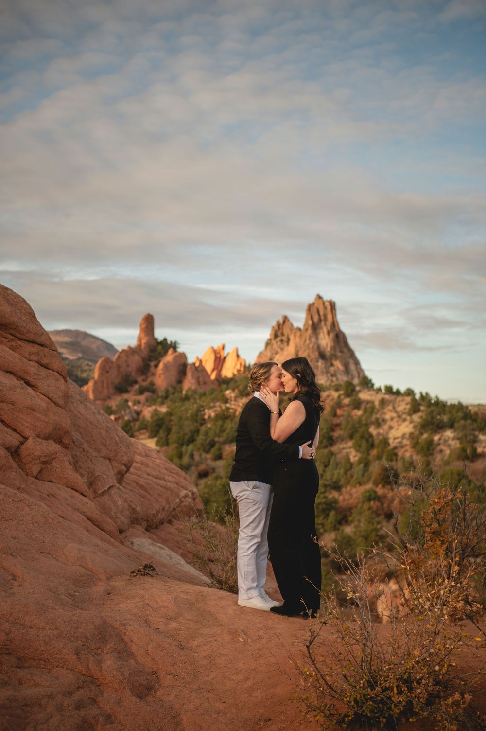 Couple embracing and kissing at Garden of the Gods, Colorado, with red rock formations and blue sky at sunset.