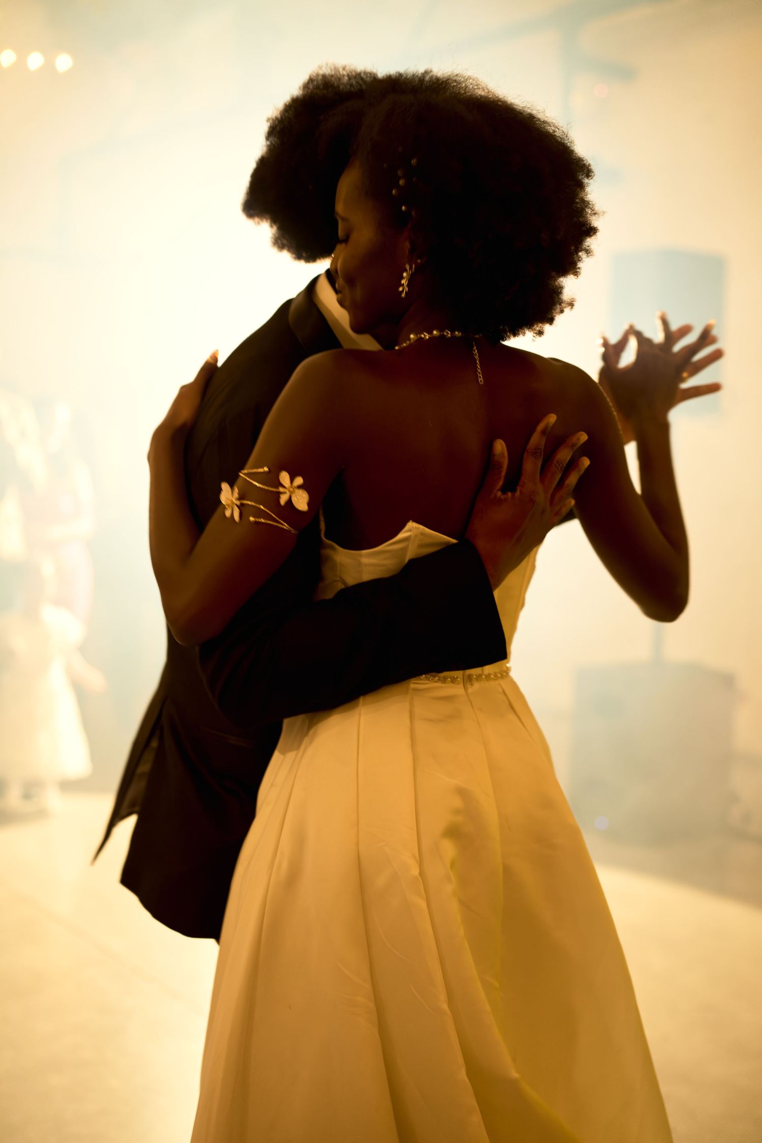 Couple dancing elegantly at a formal event, woman in white gown with floral tattoo, warm golden lighting.