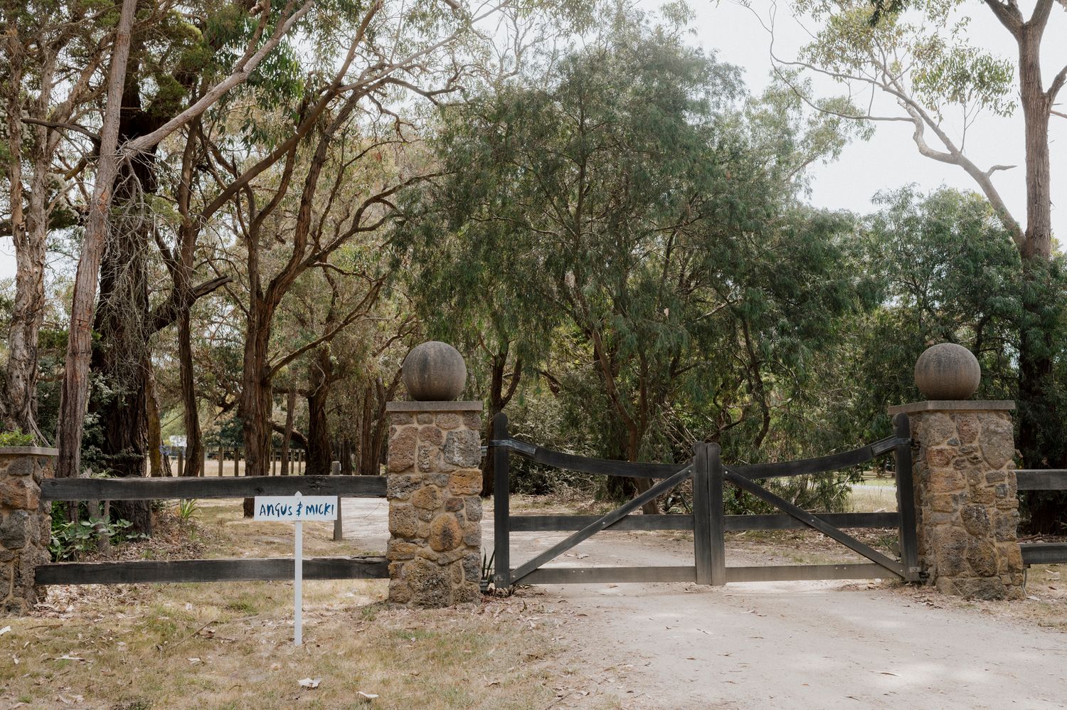 Stone pillar gate entrance with wooden gates leading to a tree-lined driveway on a rural property.