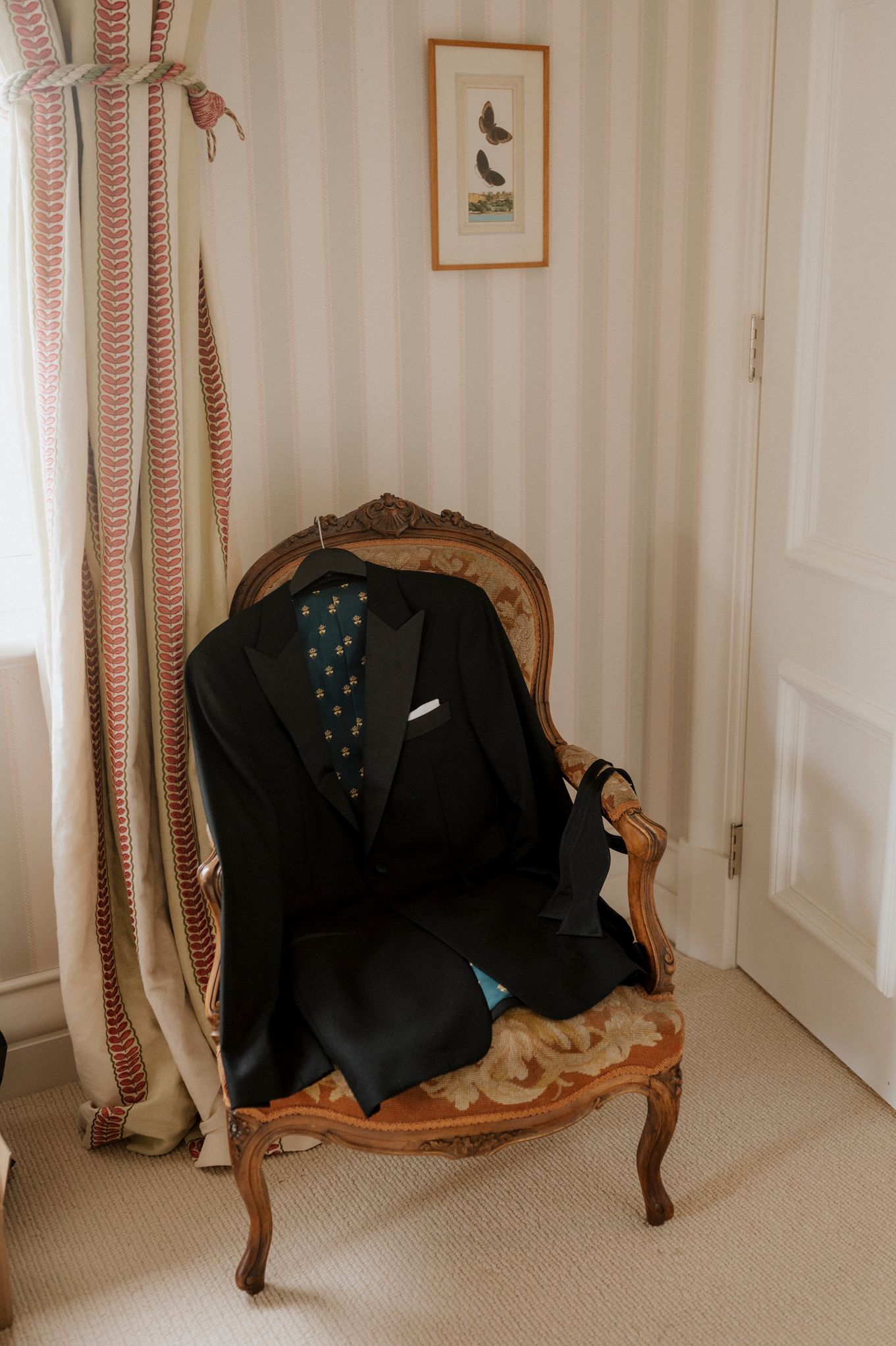Black tuxedo jacket with teal polka dot tie draped over ornate vintage French chair in elegant striped wedding suite.