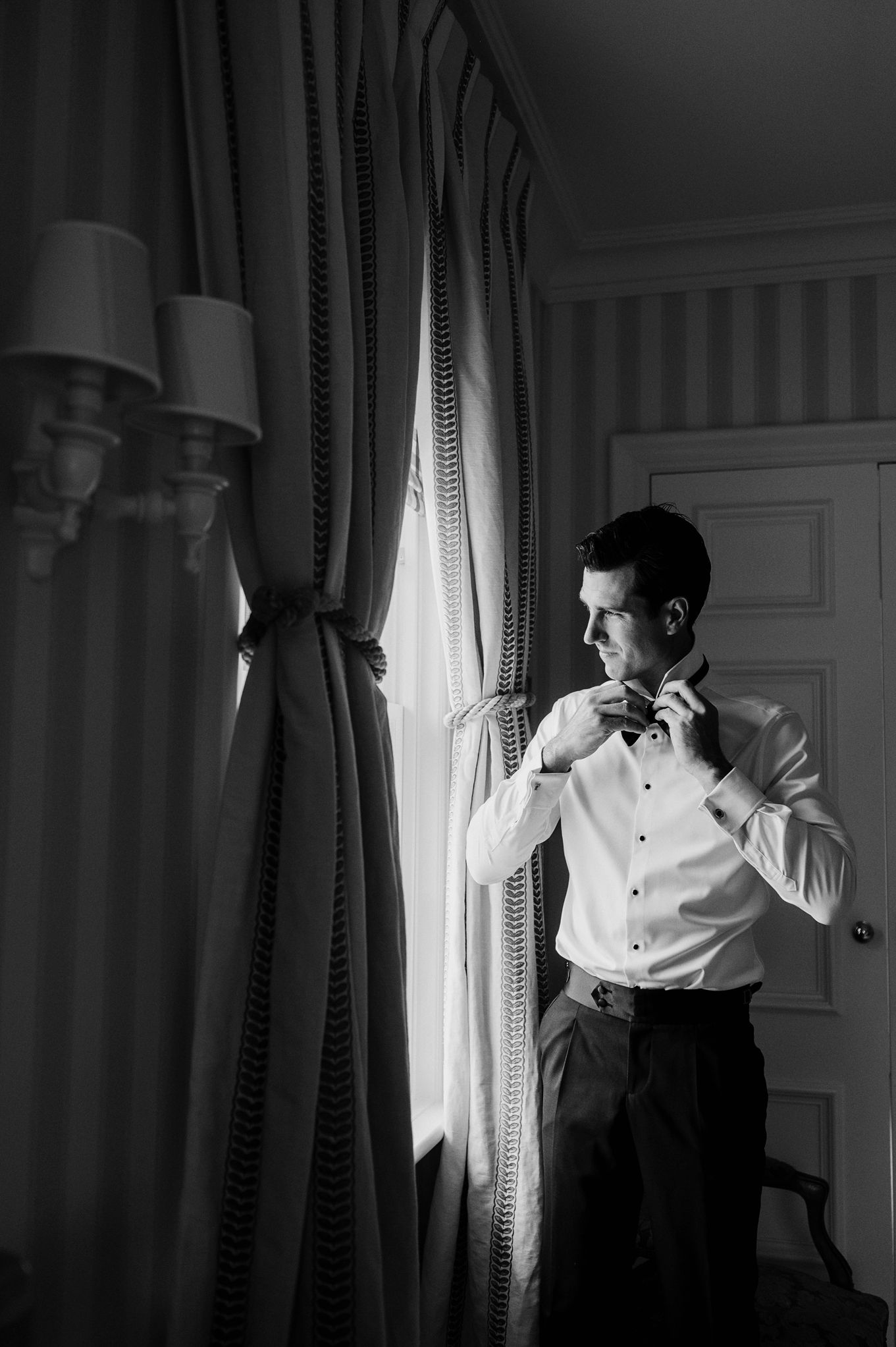 Black and white photo of a groom adjusting his bow tie by a window in elegant formal attire.